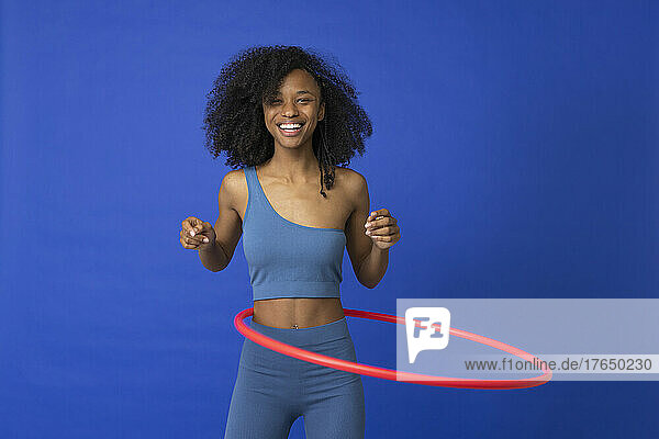 Smiling woman spinning red hoop around waist against blue background