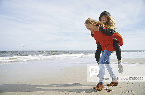 Happy blond mother giving piggyback ride to daughter at beach on sunny day