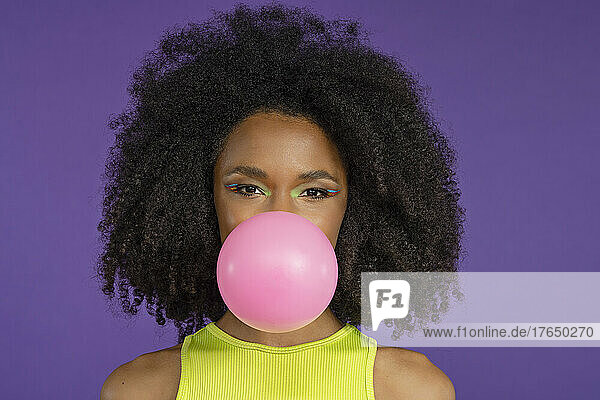 Beautiful young woman blowing pink bubble gum against purple background