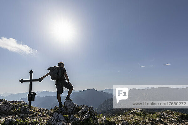 Germany  Bavaria  Sun shining over silhouette of male hiker standing by Fockenstein summit cross
