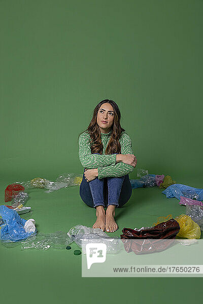 Thoughtful young woman sitting amidst plastic waste against green background