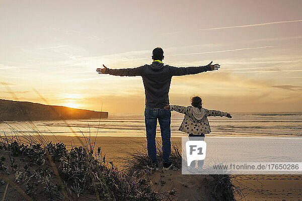 Carefree father and daughter standing on sand dune with arms outstretched at beach