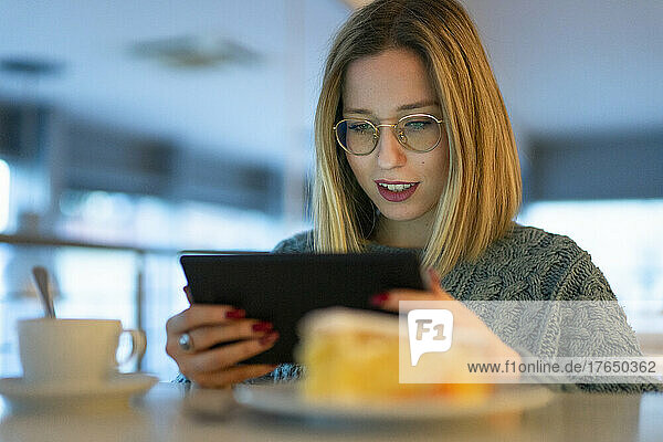 Junge Frau mit blonden Haaren sitzt am Tisch und benutzt einen Tablet-PC