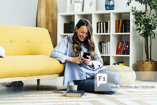 Happy woman using smart phone sitting on carpet by sofa at home