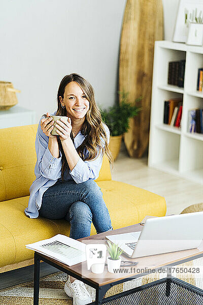 Happy freelancer holding coffee cup sitting on sofa at home
