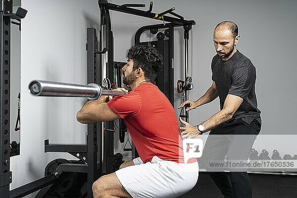 Fitness instructor giving support to young man lifting barbell at health club
