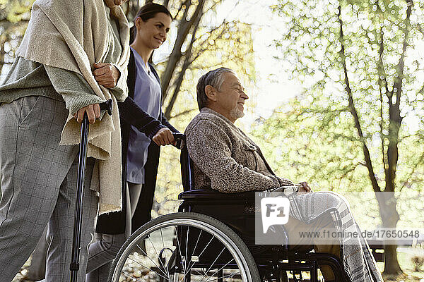 Ältere Frau geht mit Stock an Krankenschwester vorbei und behinderter Mann sitzt im Rollstuhl im Park