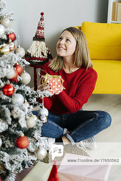Smiling woman with Christmas present looking at decorated tree in living room at home