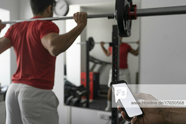 Hand of instructor holding smart phone near young man lifting barbell at gym