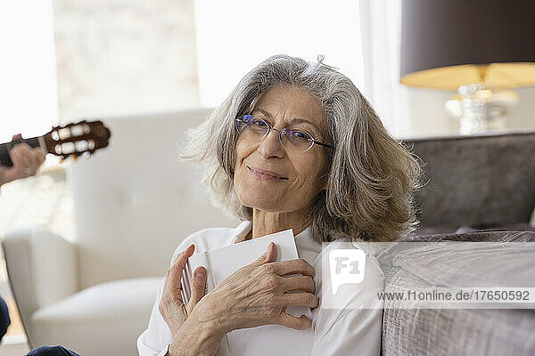 Lächelnde ältere Frau mit Brille sitzt mit Buch zu Hause auf dem Sofa
