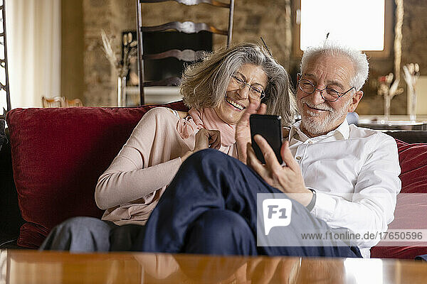 Happy senior couple having video call on mobile phone sitting on sofa at boutique hotel