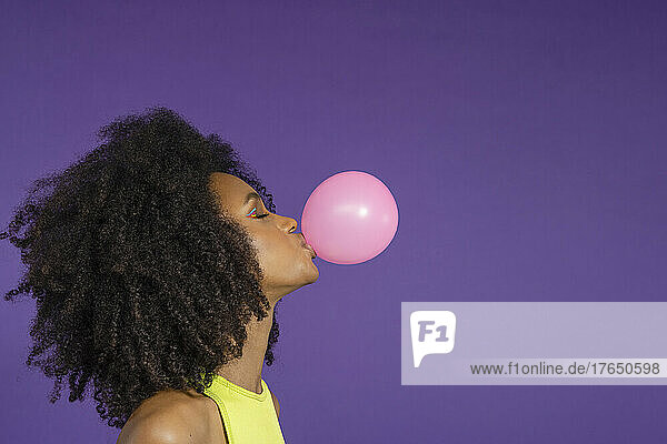 Young woman with eyes closed blowing pink bubble gum against purple background