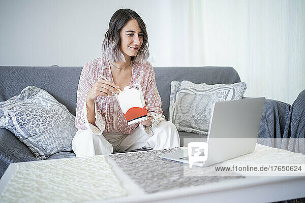 Smiling woman with take out food watching video through laptop sitting on sofa at home