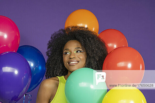 Smiling young woman with curly hair amidst colorful balloons against purple background