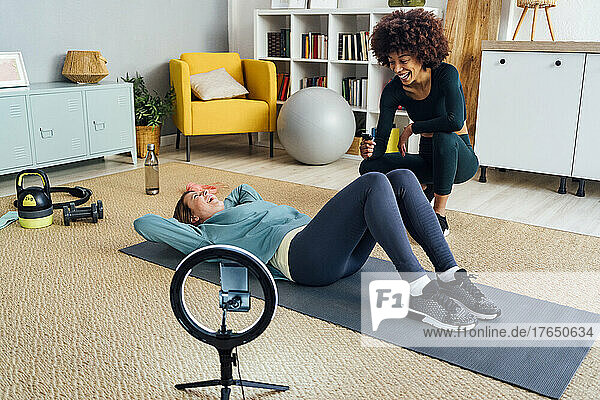 Cheerful woman crouching by friend lying with hands behind head on exercise mat in living room
