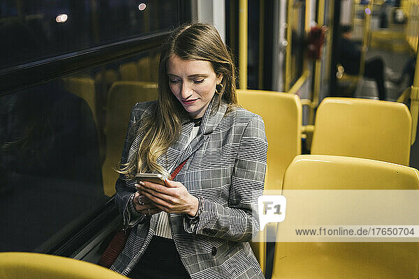 Smiling young woman using mobile phone sitting by window in tram