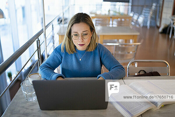 Blonde junge Frau benutzt Laptop und liest ein Buch am Tisch