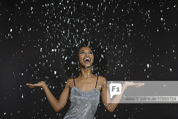 Happy young woman with mouth open standing under falling confetti against black background