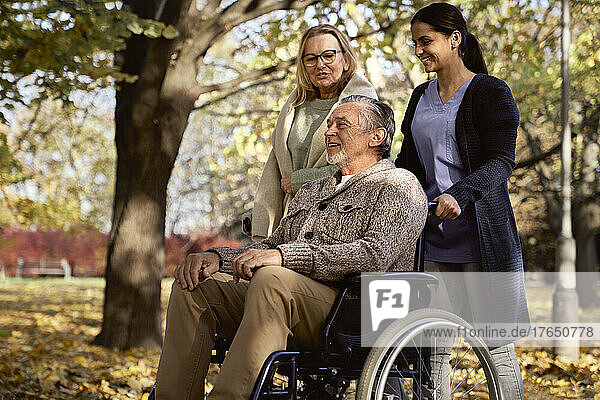 Smiling senior woman talking with man sitting in wheelchair pushed by nurse at park