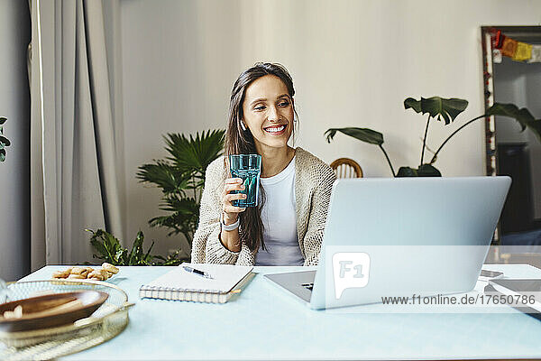 Happy freelancer holding glass of water working on laptop at home