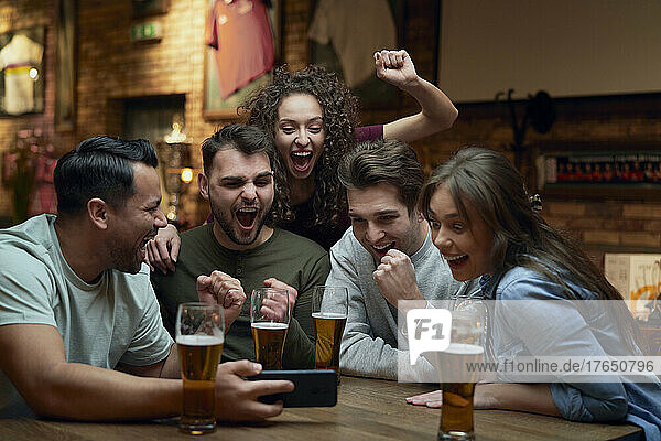 Gruppe fröhlicher Fußballfans  die in einer Kneipe Bier trinken und sich ein Spiel auf dem Smartphone ansehen