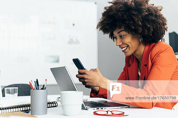 Happy businesswoman using smart phone sitting with laptop at desk in office