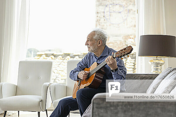 Senior man playing guitar sitting on sofa in living room at home