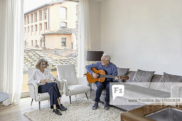 Senior man playing guitar looking at woman reading book in living room