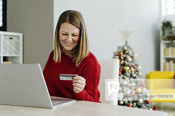 Smiling woman looking at credit card sitting with laptop in living room