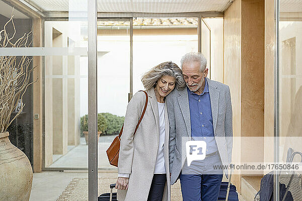 Smiling senior couple walking with suitcases in lobby at boutique hotel
