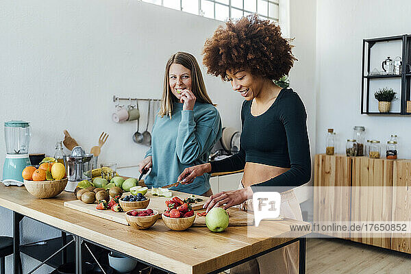 Smiling young woman cutting strawberries by friend eating apple at table in kitchen