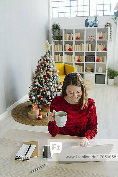 Happy woman with laptop and coffee cup sitting in living room at home