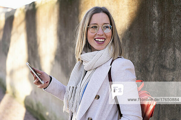 Lächelnde junge Frau mit Mobiltelefon vor der Wand