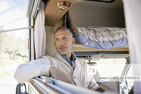 Thoughtful man with book looking through window of camper van