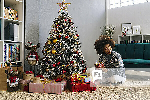 Happy woman sitting with gifts by Christmas tree at home