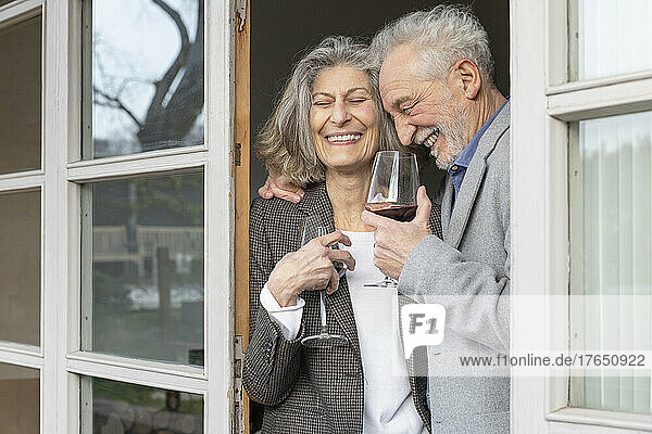 Cheerful senior couple with wineglasses standing at doorway