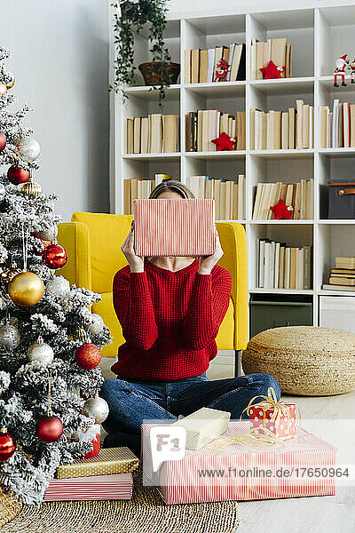 Lächelnde Frau bedeckt Gesicht mit Weihnachtsgeschenk im heimischen Wohnzimmer