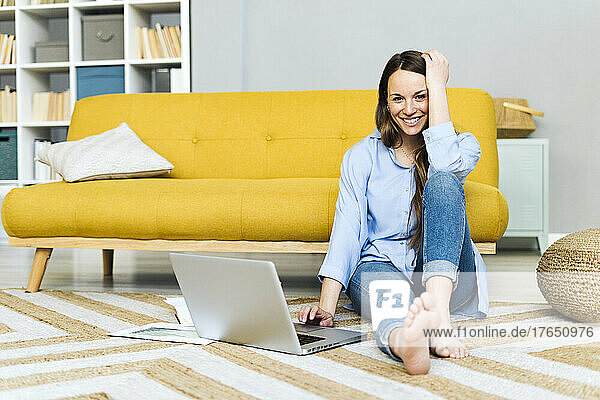 Happy freelancer with laptop sitting on carpet by sofa at home
