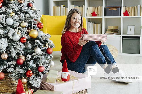 Glückliche Frau mit Geschenk sitzt zu Hause am Weihnachtsbaum