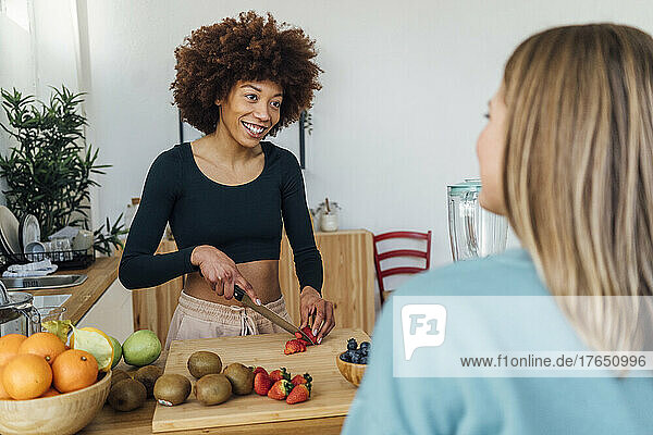 Happy young woman cutting strawberries talking with friend in kitchen