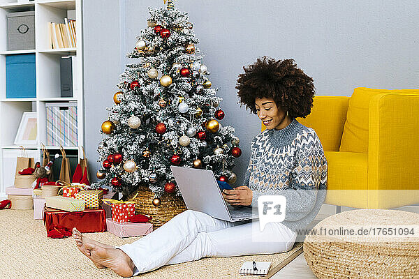 Smiling woman with laptop using smart phone by yellow armchair at home