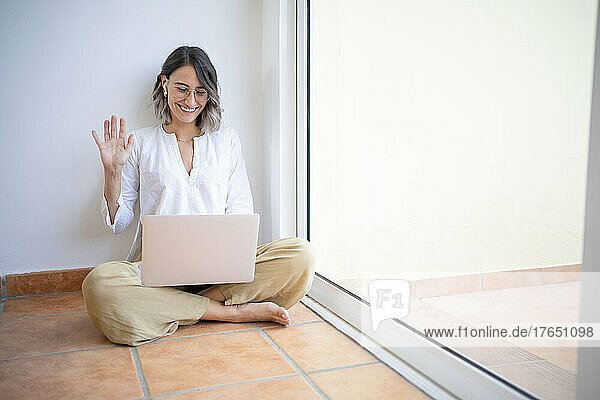 Happy freelancer waving hand on video call through laptop sitting cross-legged by window