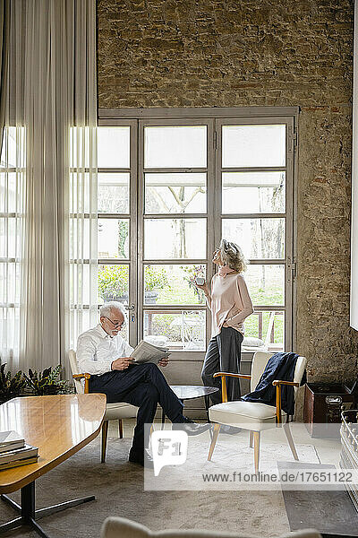 Woman holding coffee cup standing with hand in pocket by man reading newspaper in living room at home