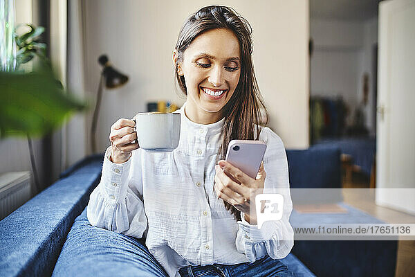 Lächelnde Frau mit Kaffeetasse und Smartphone  die zu Hause auf dem Sofa sitzt