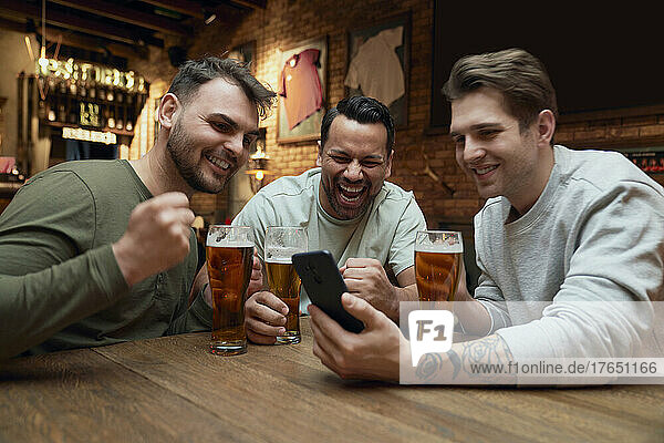 Three cheerful male soccer fans having beer and watching a match on smartphone in a pub