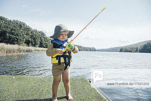 Cute baby boy with fishing rod standing by river on sunny day
