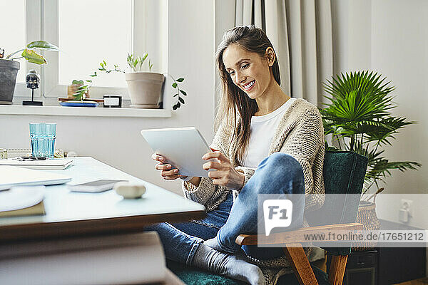 Lächelnde Frau  die zu Hause auf einem Stuhl sitzt und einen Tablet-PC benutzt