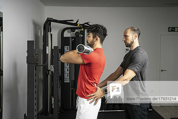 Instructor giving support to young man lifting barbell at gym