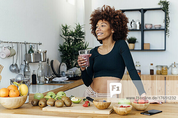 Smiling woman with Afro hairstyle holding glass of smoothie standing in kitchen