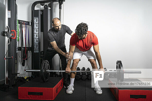 Coach giving support to young man lifting barbell at health club